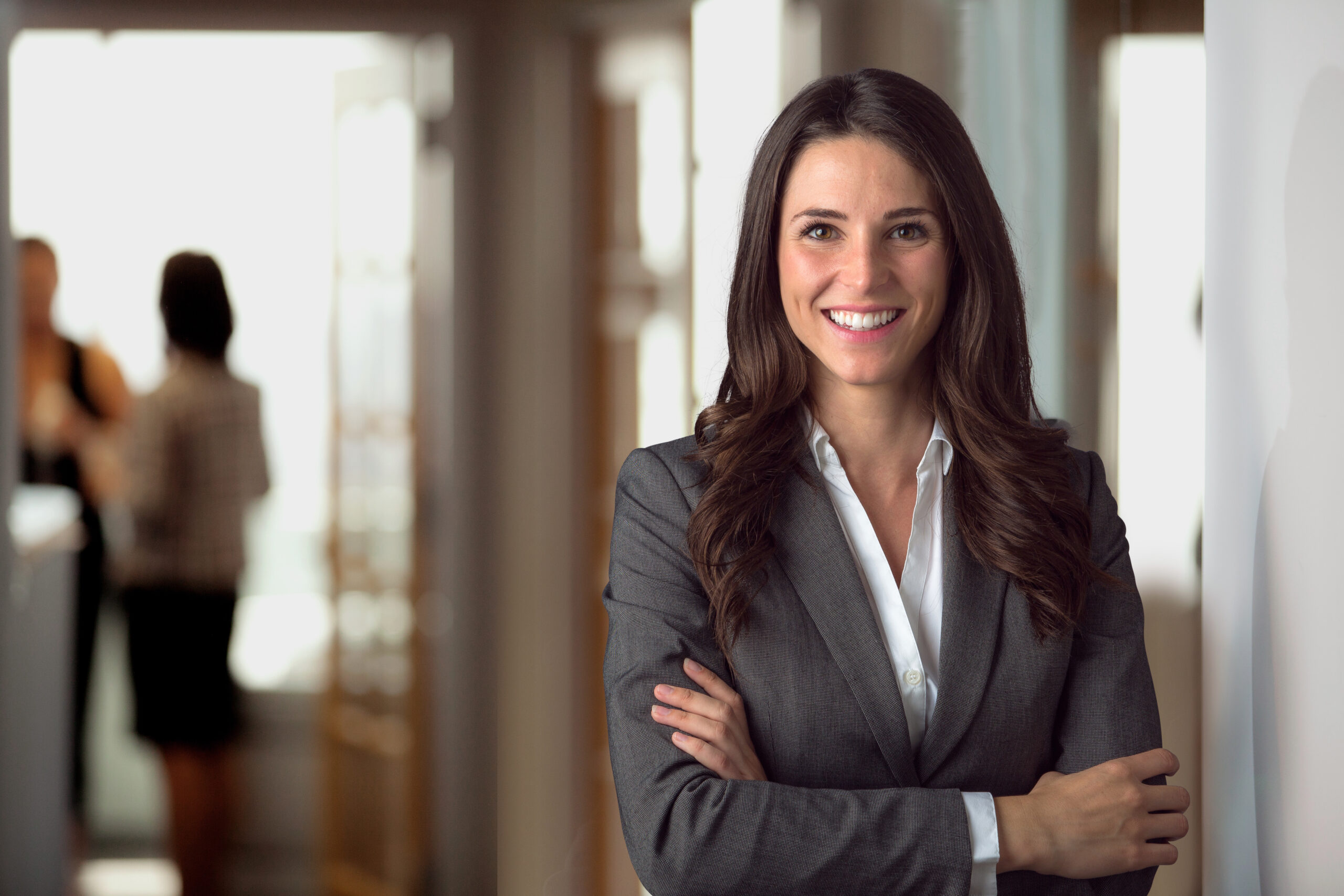 A professional businesswoman smiling while standing with arms crossed in an office environment.