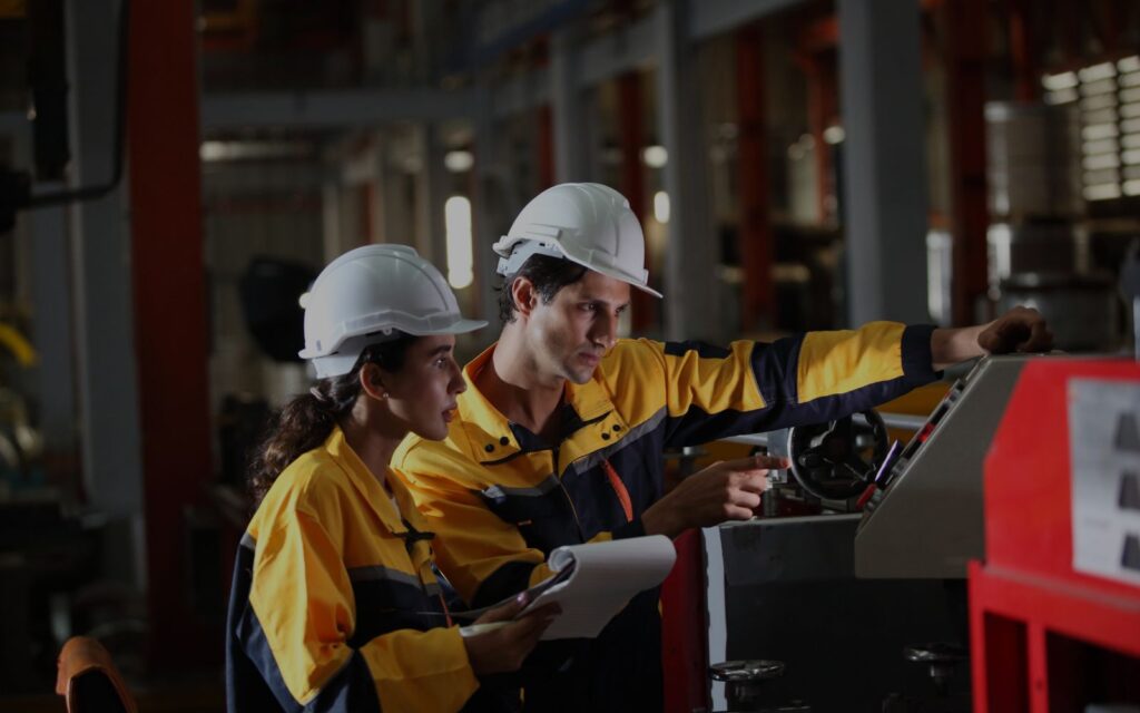 Two engineers in safety gear inspecting a machine in a manufacturing facility. GMPPros logo in the top left corner, emphasizing expertise in industrial solutions.