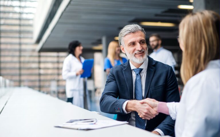 A businessman in a suit shakes hands with a person in a lab coat in a modern, bright office setting, with other medical professionals in the background.