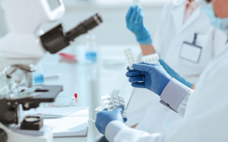 Scientists in lab coats handling pill packs with a microscope and lab equipment in the background, focusing on pharmaceutical research.