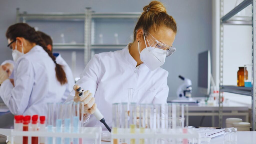 Batch Record Errors in Pharma: Types, Causes & Prevention Strategies 5 Scientist in lab coat, safety goggles, and mask using a pipette to transfer liquid into test tubes in a laboratory setting.