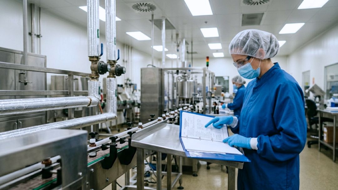 Operator in cleanroom gear reviewing batch record binder near production line, illustrating in-process control documentation failures and prevention.
